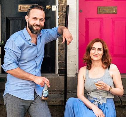 The husband and wife team from Cambucha sit on Cambridge house steps while drinking Kombucha and smiling
